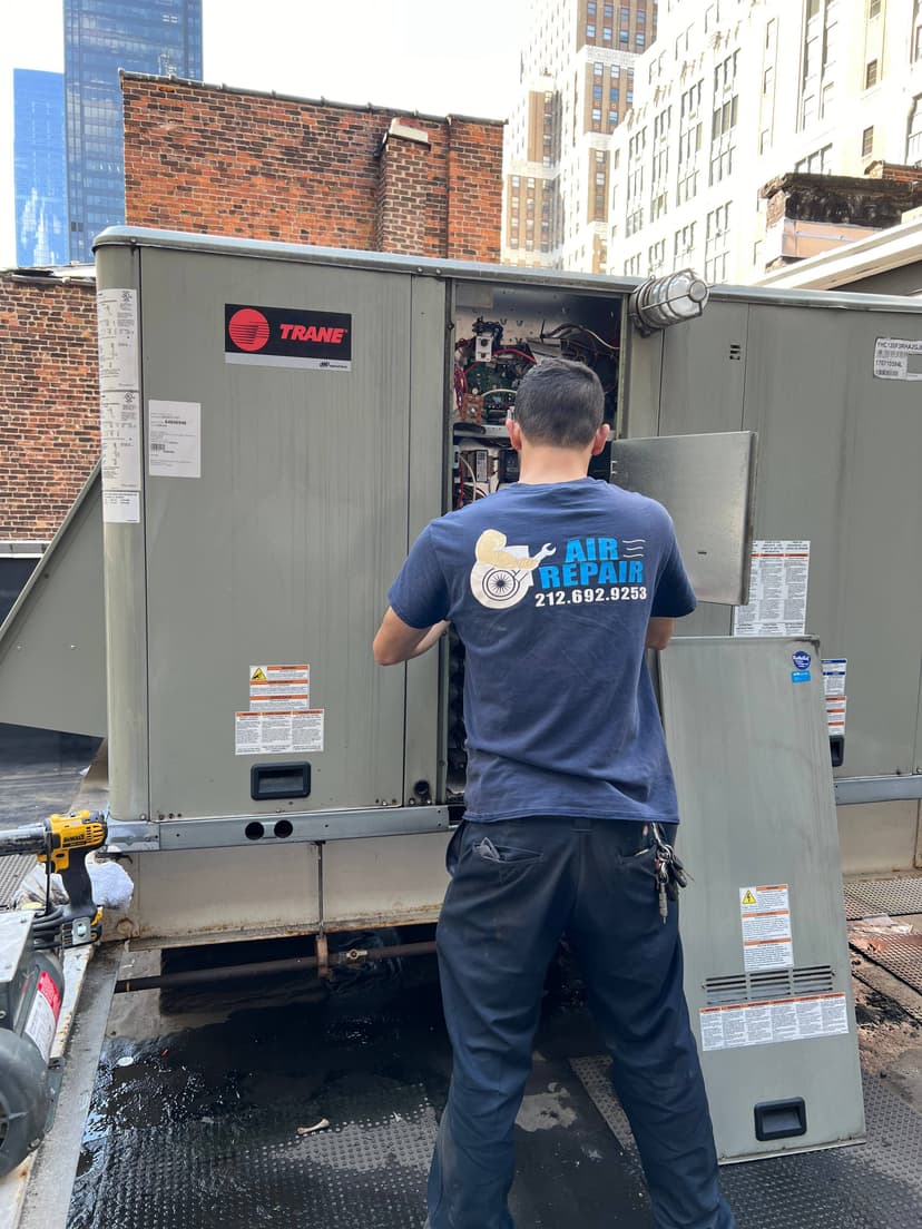 A technician in a blue Air Repair shirt, proudly displaying the company's branding, works on an open HVAC unit on a rooftop in an urban area.