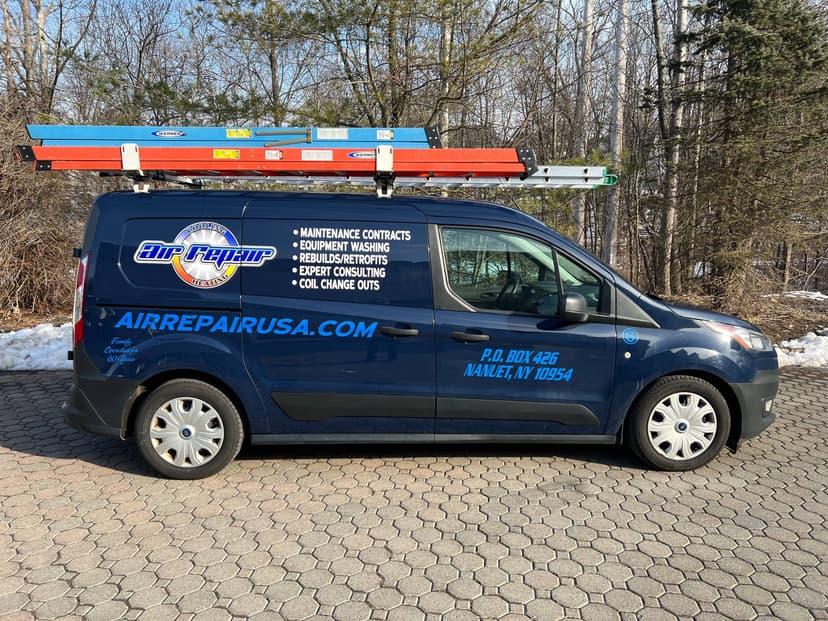 A blue Air Repair LLC work van is parked on a paved driveway with a wooded area in the background. The van, equipped with ladders on the roof, proudly displays contact information and service details on the side.