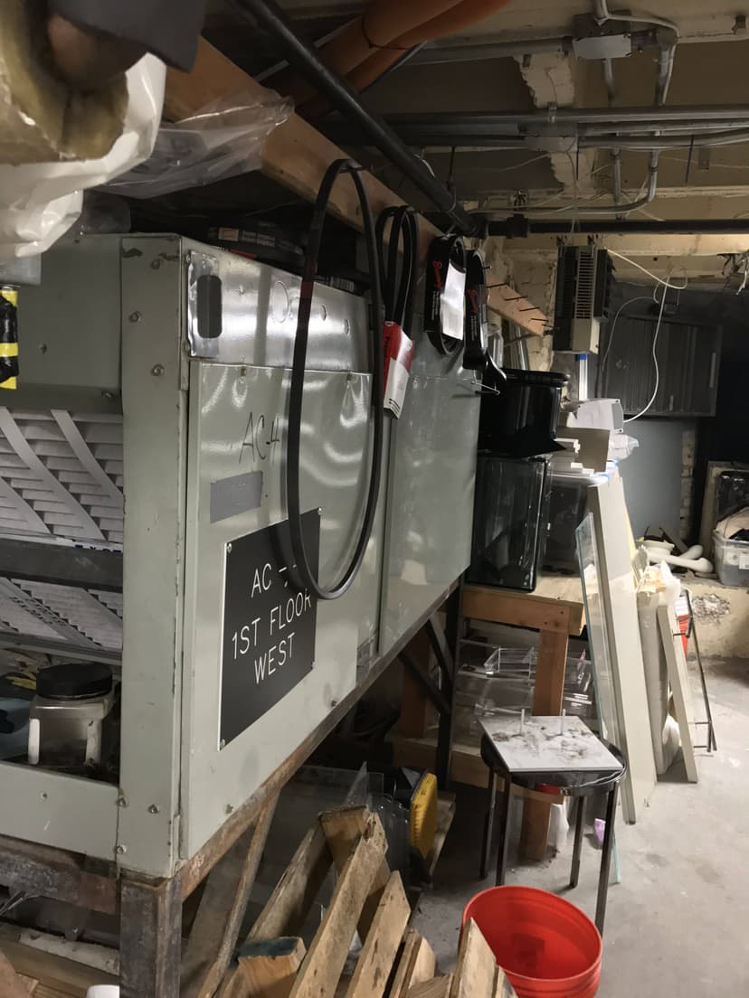 An old air conditioning unit marked 'AC - 1st FLOOR WEST' sits amid various tools and pieces of HVAC equipment in a cluttered basement.