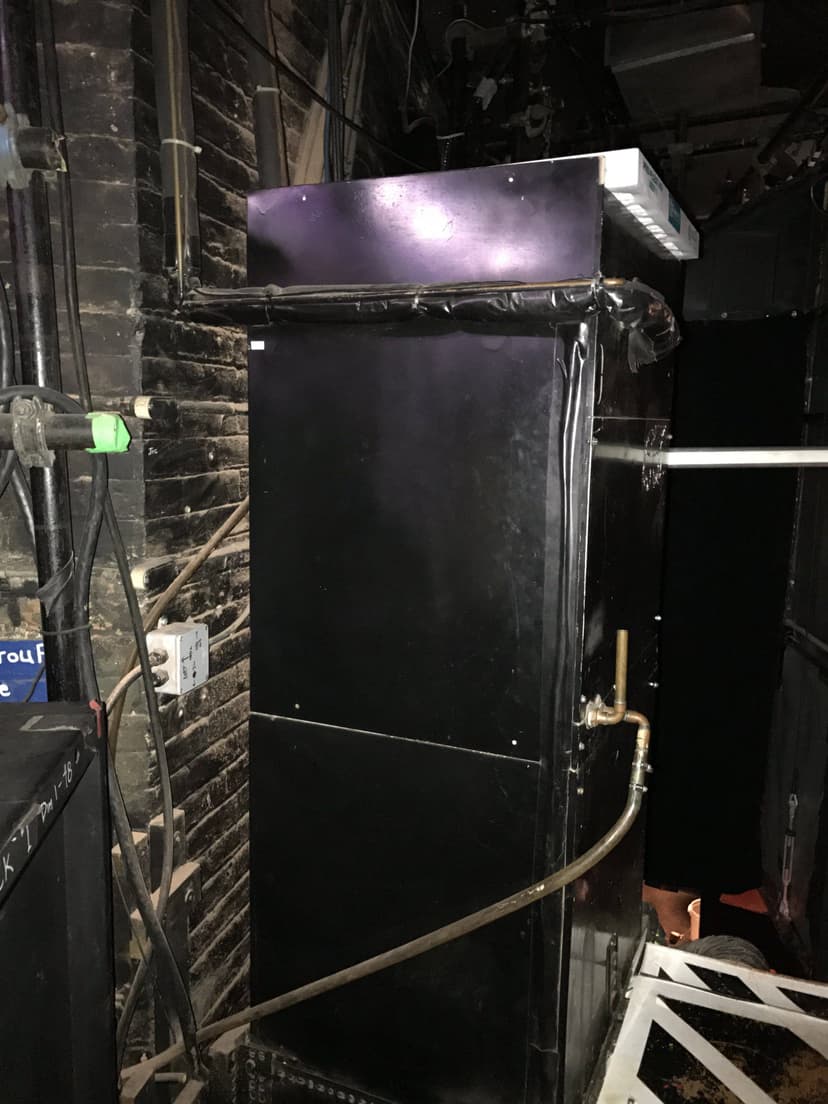 A large black industrial machine, resembling an air handler HVAC unit, stands with pipes extending from it, situated in a dimly lit area with brick walls.