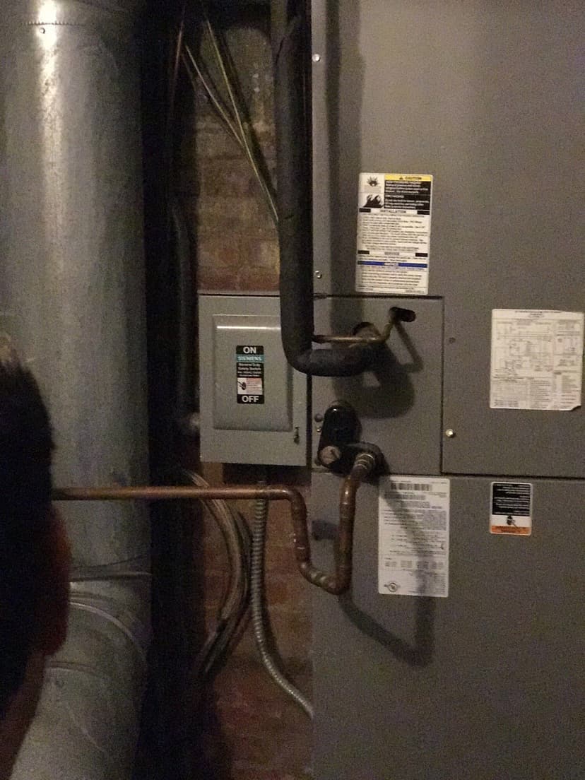 A person stands in front of an air handler and furnace in a dimly lit basement, inspecting the HVAC equipment.