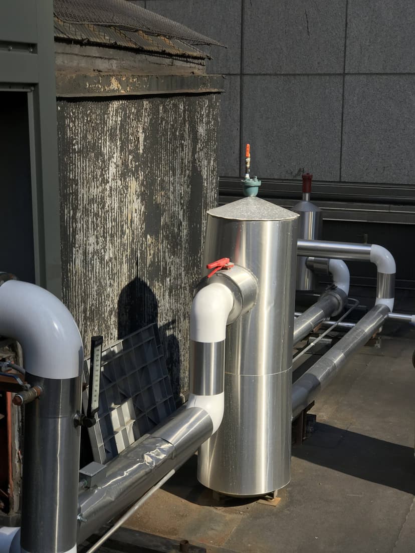 Industrial pipes and a chiller HVAC unit stand alongside metallic structures against a weathered building wall on the rooftop.