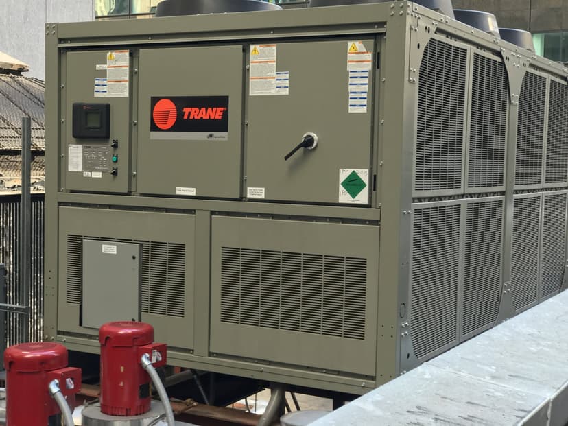 Large industrial chiller HVAC unit with a Trane logo installed outdoors, surrounded by a fence and pipes in an urban setting.