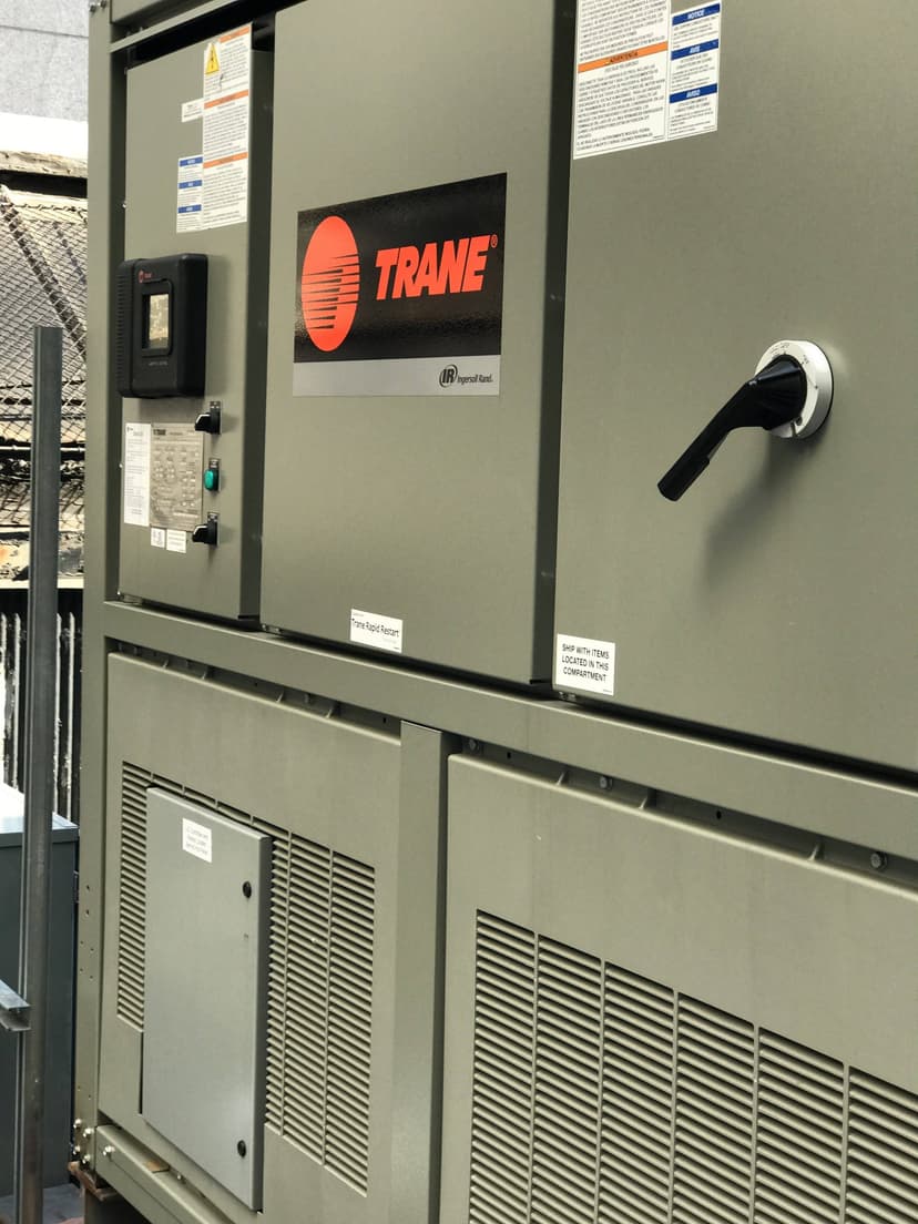 Close-up of a Trane Chiller HVAC unit with control panel and labels, located outdoors near a metal fence, serving as an industrial example of modern cooling technology.