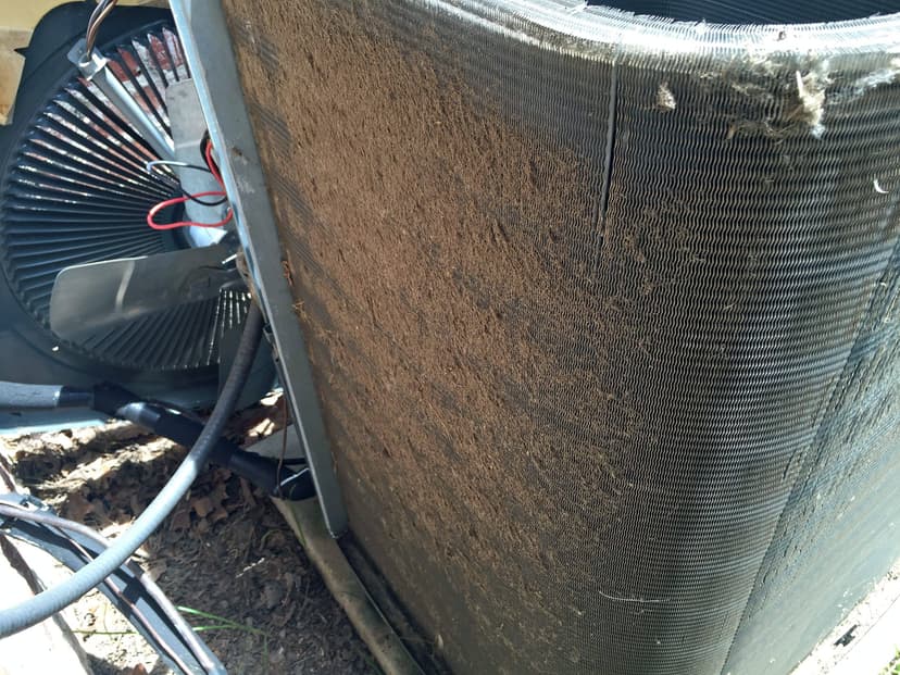 A dirty outdoor HVAC condenser, an example of an HVAC unit coil, is covered in dust and debris, sitting next to a fan with visible wires.