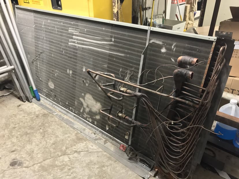 A large metal air conditioning condenser unit with connected copper tubing and wires, ideal for a workshop, features an efficient coil in cooling.