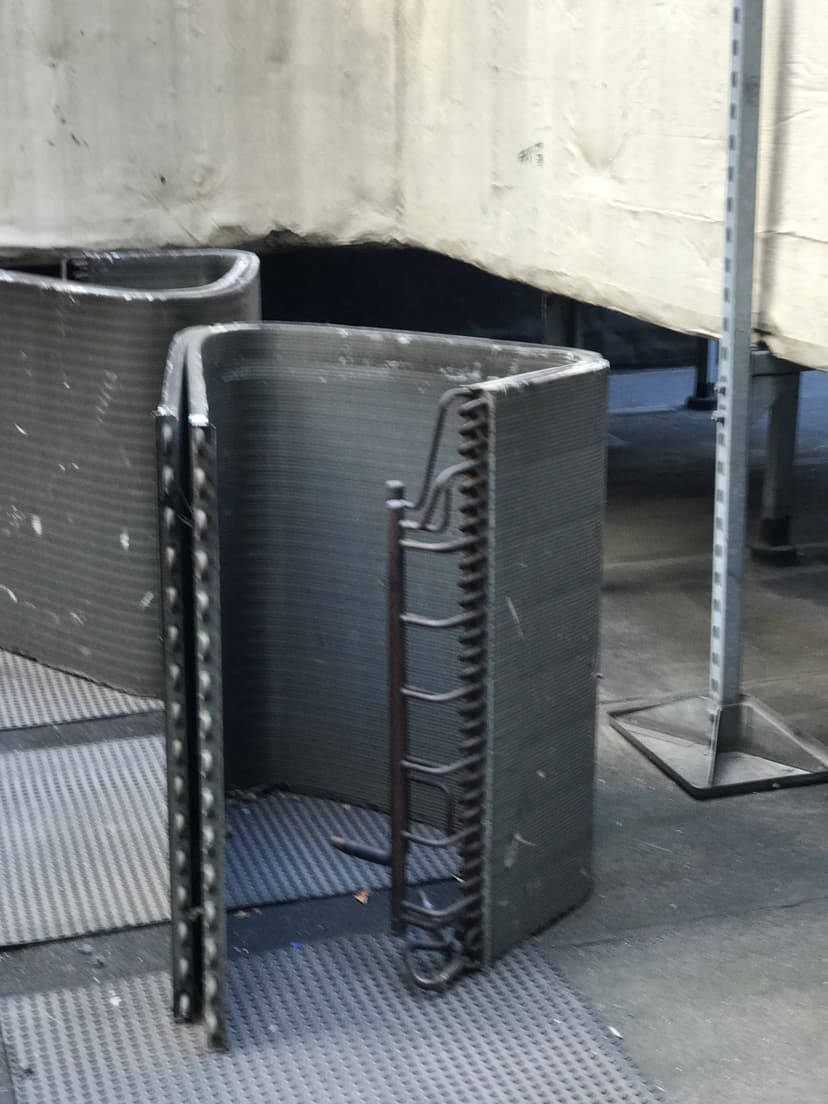 A curved, gray, industrial piece of equipment with a ladder beside it, sitting on a textured mat, featuring a coil for cooling or heating unit replacement.