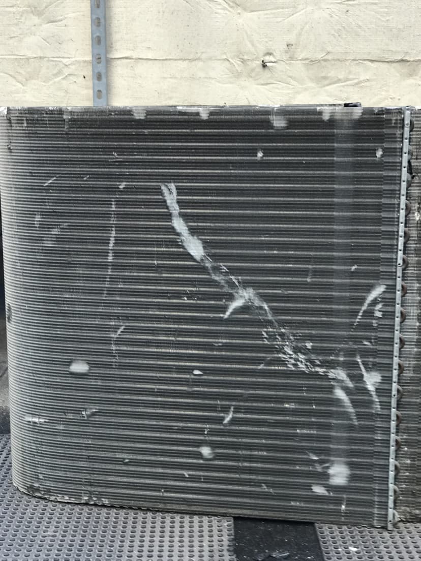 A damaged air conditioning condenser coil with visible dents and spots, positioned against a wall, highlighting the need for cooling unit replacement.