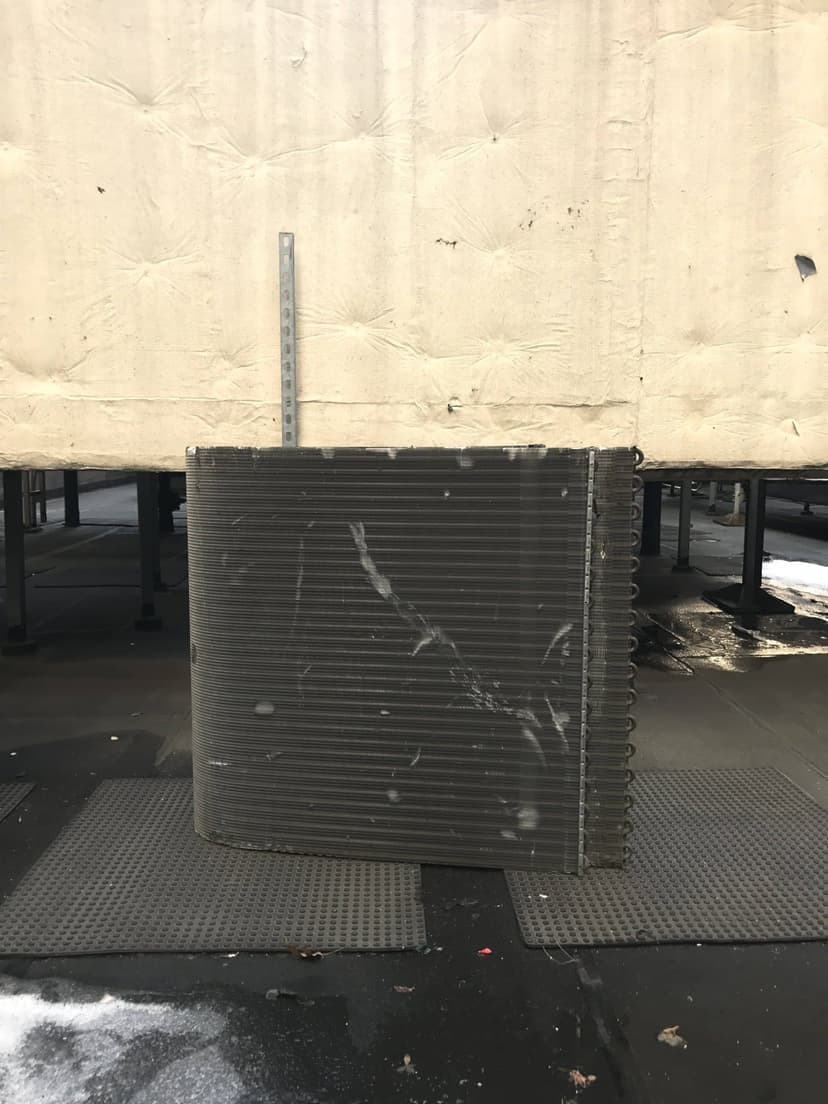 A large, coiled heat exchanger for a cooling unit replacement is placed on a rubber mat beside a beige wall on an industrial rooftop.