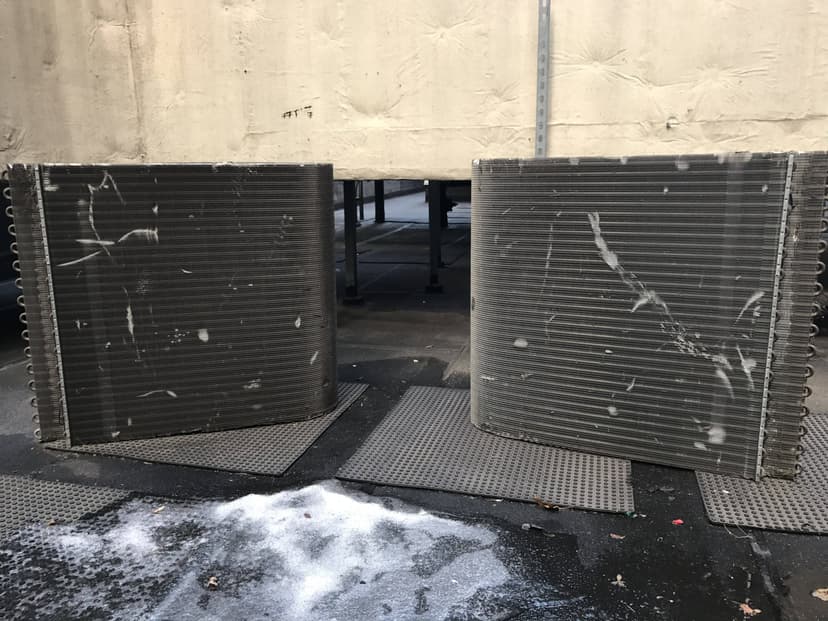 Two large, curved metal coils for a heating unit replacement stand on mats outdoors, near a structure, with some snow and dirt visible on the ground.