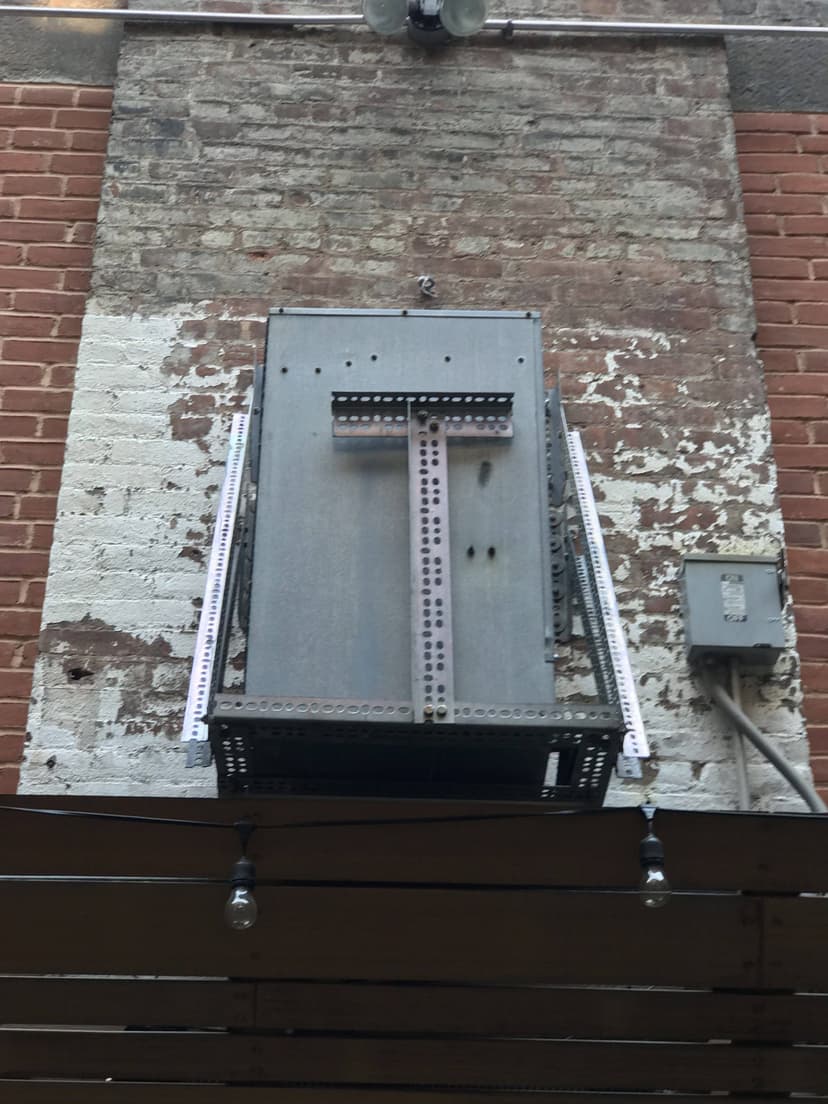 A metal structure is mounted on a brick wall with peeling paint, and a single string of hanging bulbs is visible underneath, reminiscent of the placement of condensing units.