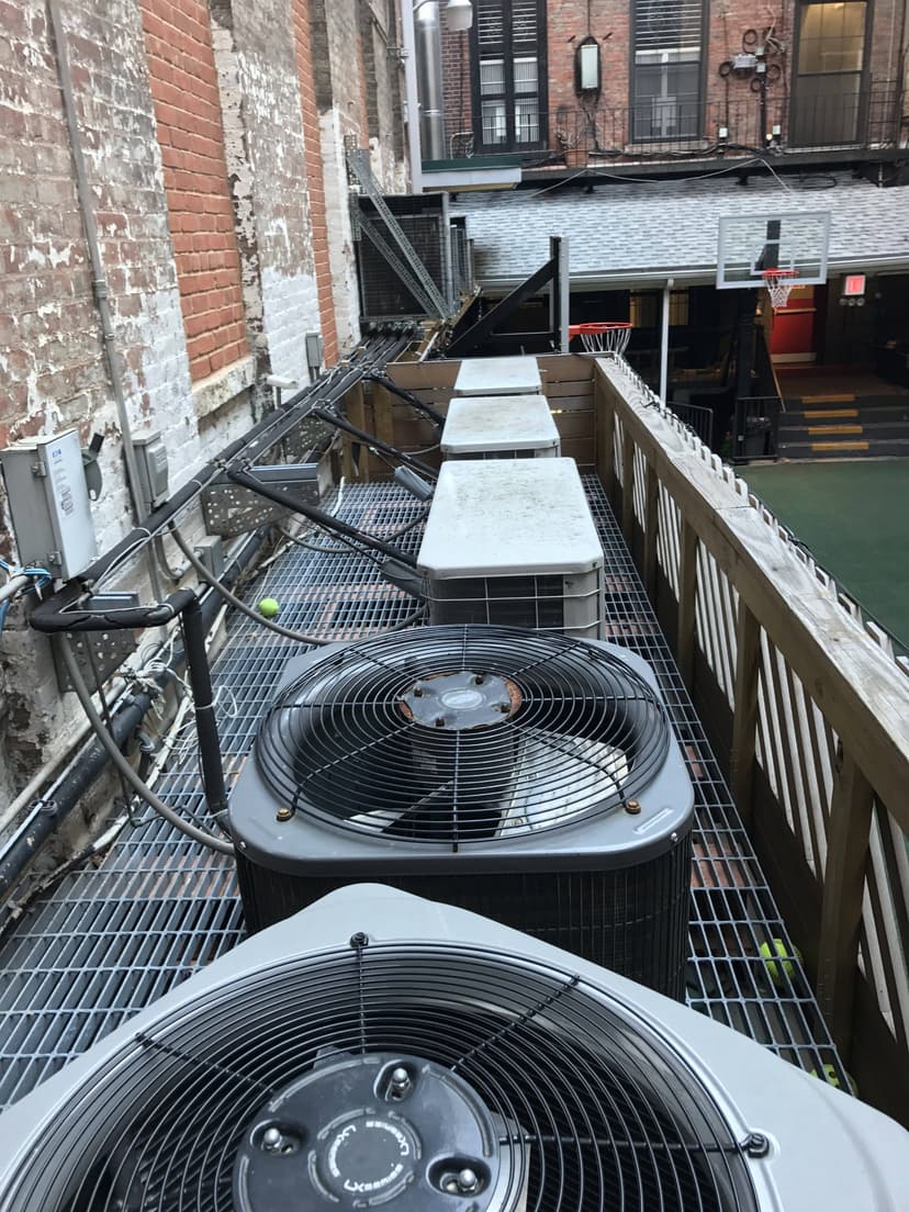 Outdoor condensing units sit on a grated platform along a brick building, with a basketball court visible in the background.