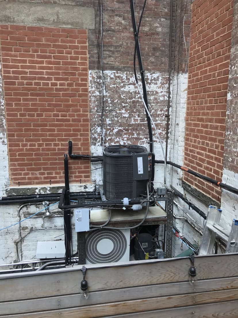 Air conditioning units and condensing units, along with electrical equipment, are mounted on a metal frame against a brick wall in an urban alleyway.