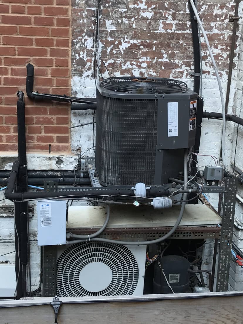 Rooftop industrial HVAC units with various pipes and wires, including condensing units, mounted against a brick and whitewashed wall.