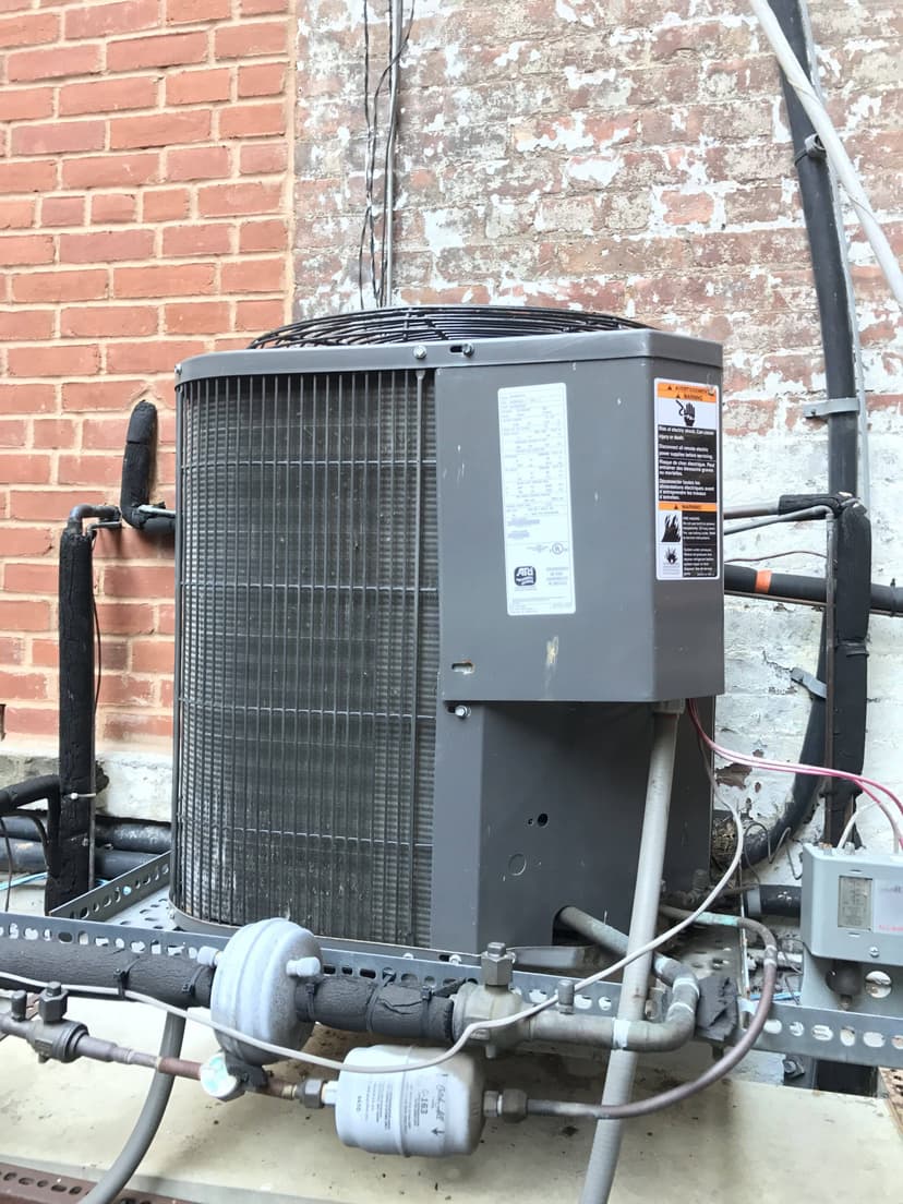 Outdoor condensing unit mounted on a brick wall with various pipes and cables attached.