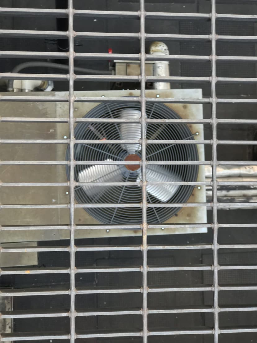 Overhead view of a metal fan and pipes, along with condensing units, seen through a grated surface.