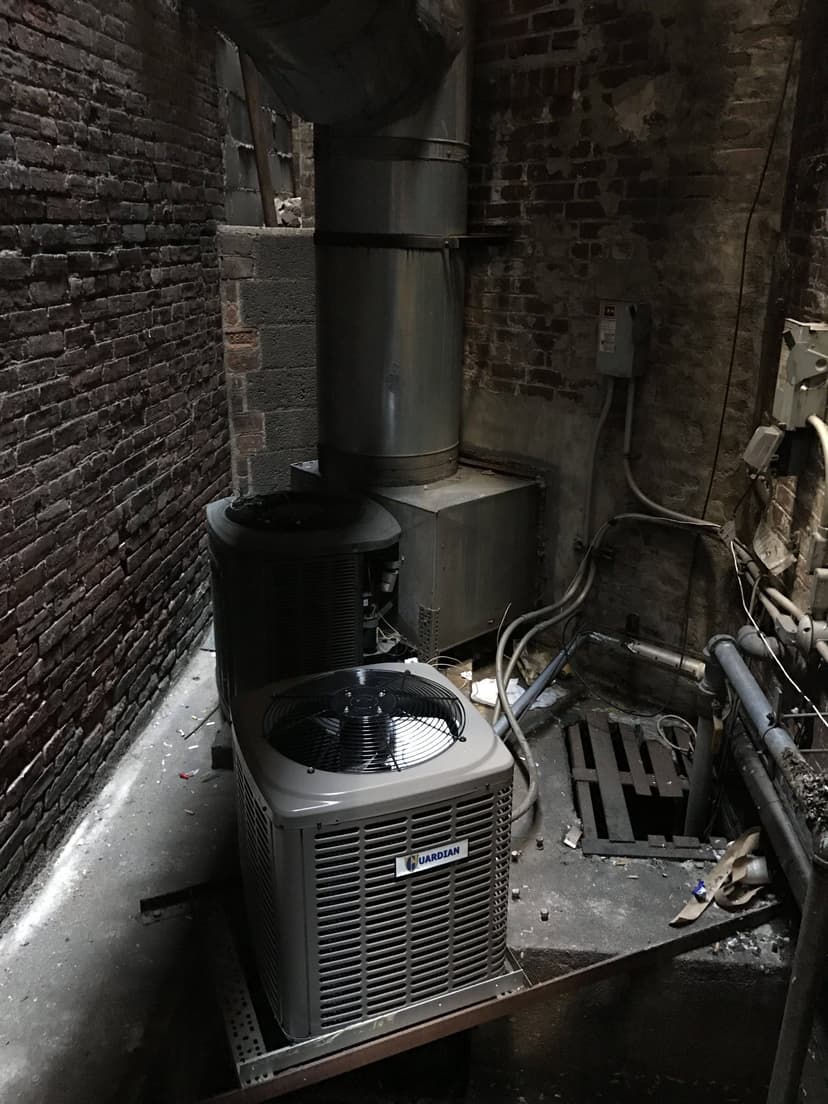 Two industrial air conditioning condensing units in a dimly lit, enclosed brick space with metal ductwork and electrical wiring.