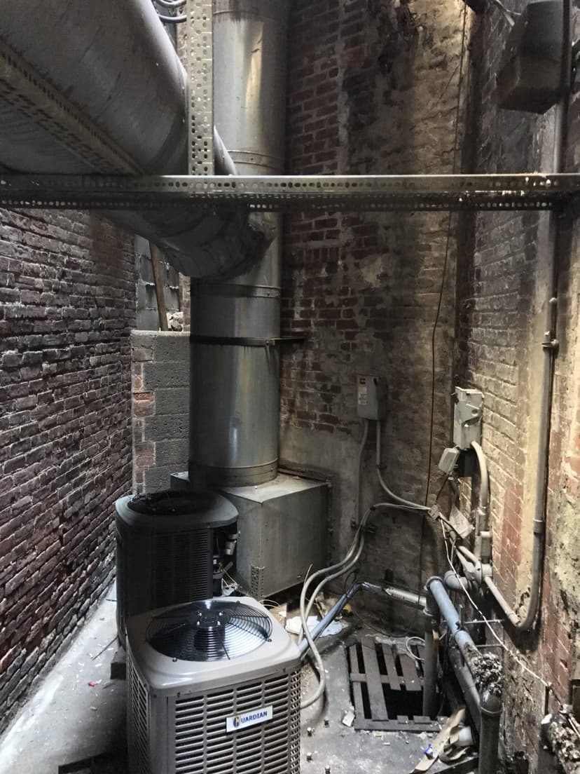 Narrow alleyway with HVAC units and condensing units, large ductwork overhead, and exposed brick walls on both sides.