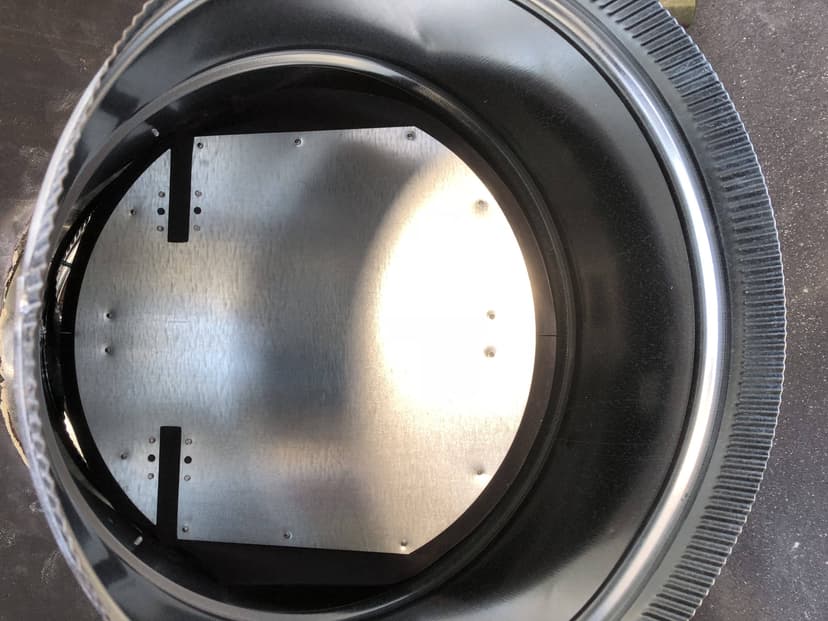 A view inside an industrial metal pipe or cylinder reveals a flat metal panel, resembling dampers for HVAC systems, attached to the interior.