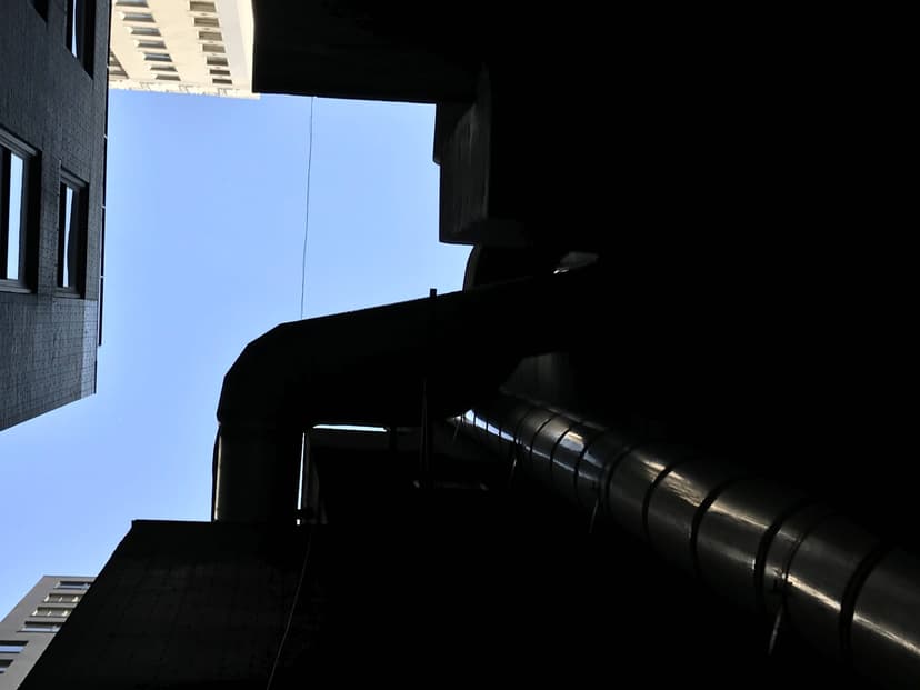 View between two city buildings with a large industrial pipe, an impressive example of HVAC duct work, running vertically across the gap under a clear blue sky.