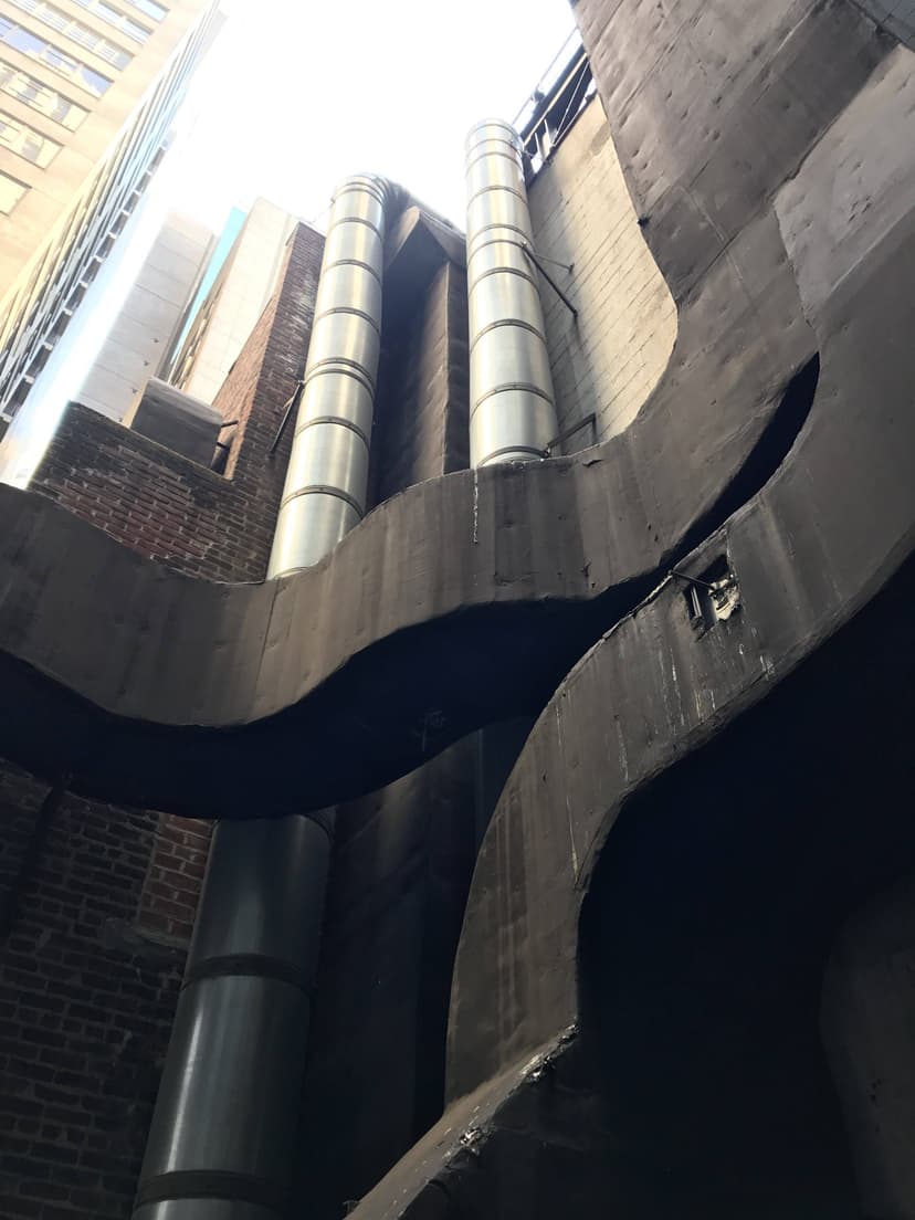 View of an alley with exposed HVAC ductwork and curved concrete structures set between tall brick and glass buildings.