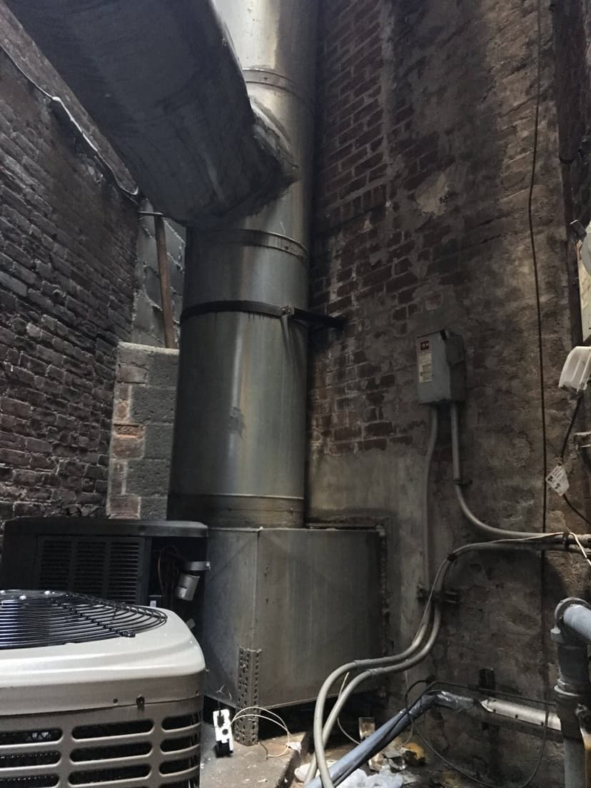 An HVAC air conditioning unit and ventilation ducts serve as a duct work example in a narrow, rustic brick and concrete area, complete with some exposed wiring.