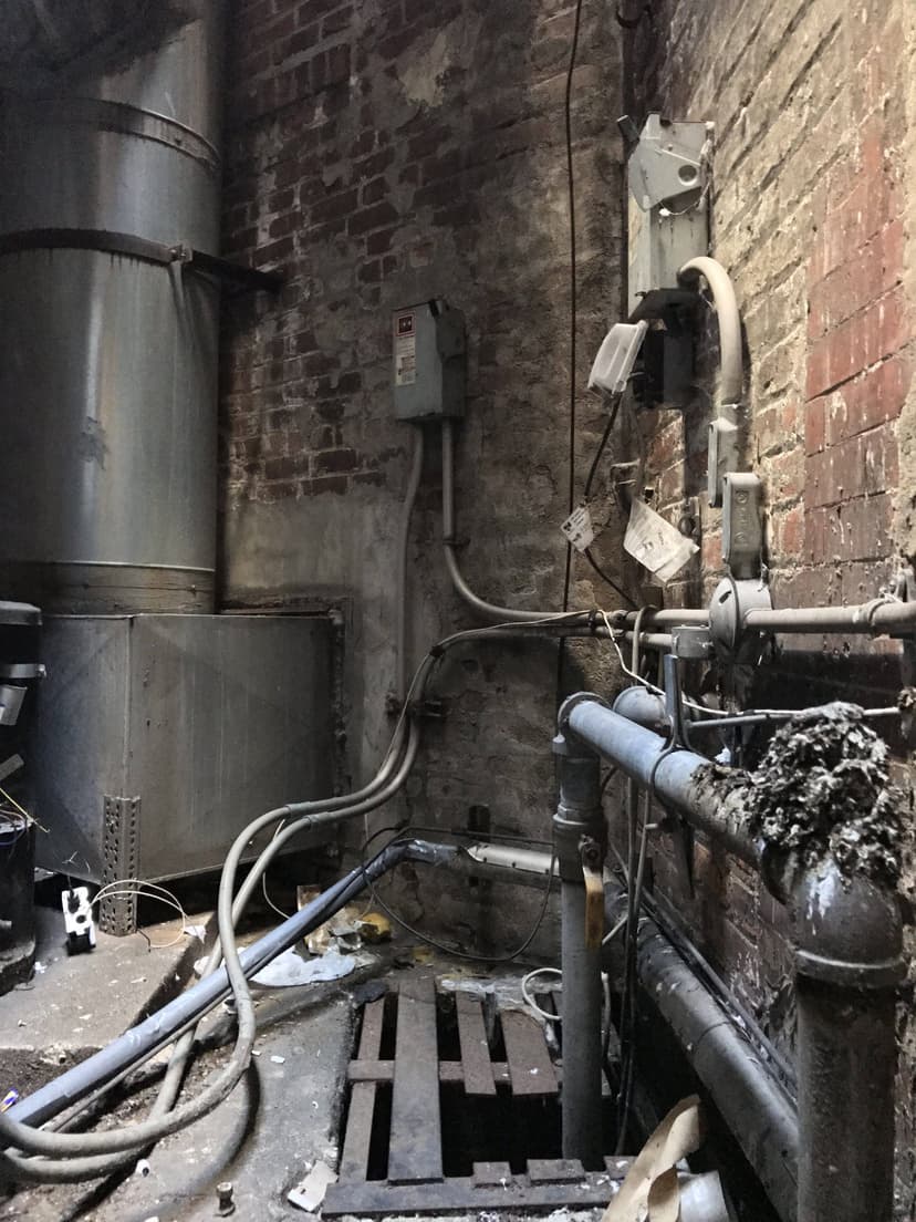 Dimly lit industrial basement with pipes, electrical panels, and a grated floor in a worn brick-walled space, serving as a classic example of HVAC duct work.