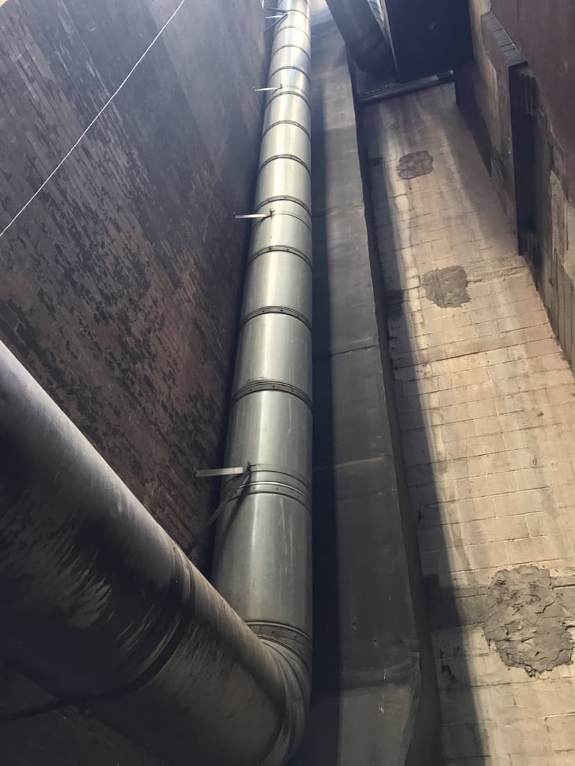 A large metal pipe, serving as an HVAC duct, runs vertically up the exterior of a tall, narrow brick and concrete building.