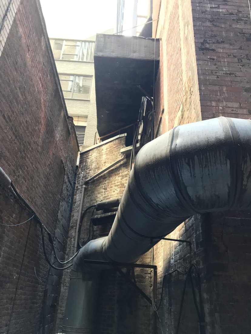 Narrow alleyway with exposed pipes, brick walls, and intricate HVAC duct work extending between buildings.