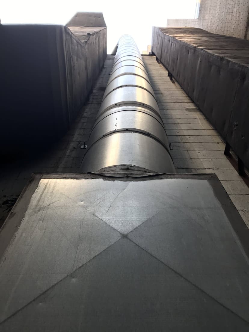 A view looking up at a metal ventilation duct, an exemplary piece of HVAC duct work, running between two tall, narrow buildings with a bright sky above.