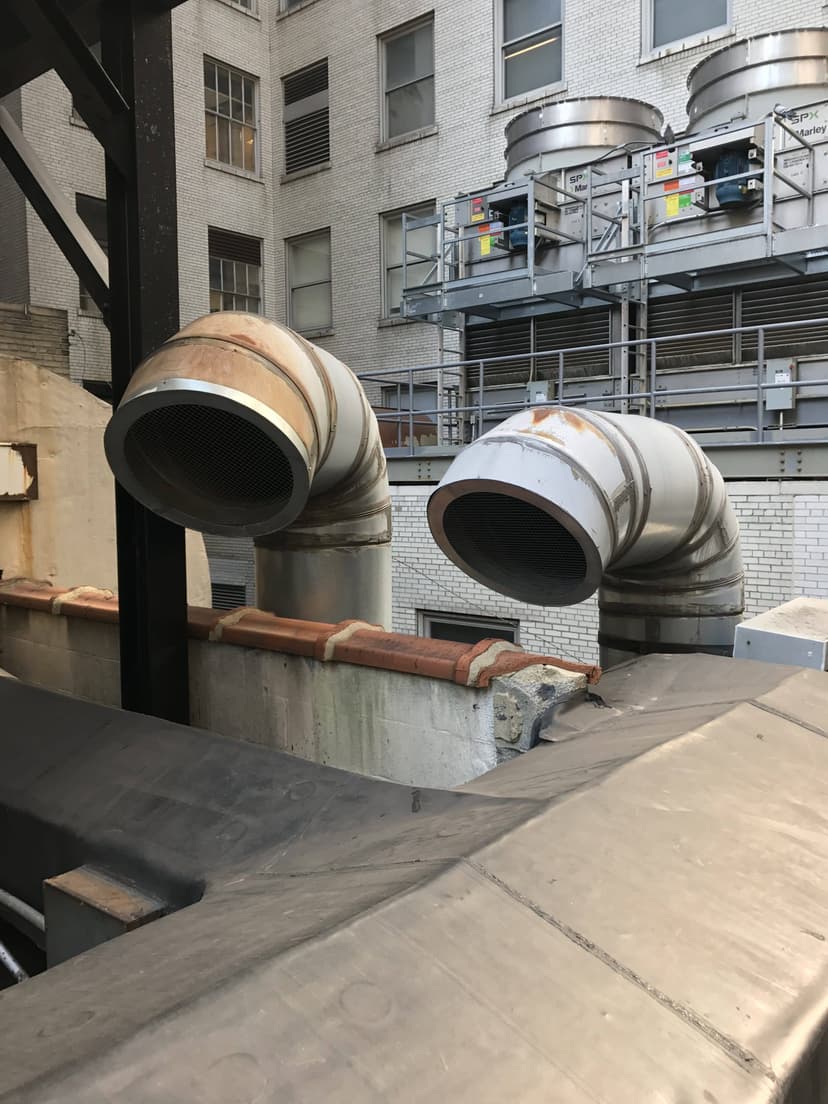 Two large industrial air ducts, a prime duct work example in modern HVAC systems, extend from a building surrounded by other urban structures.