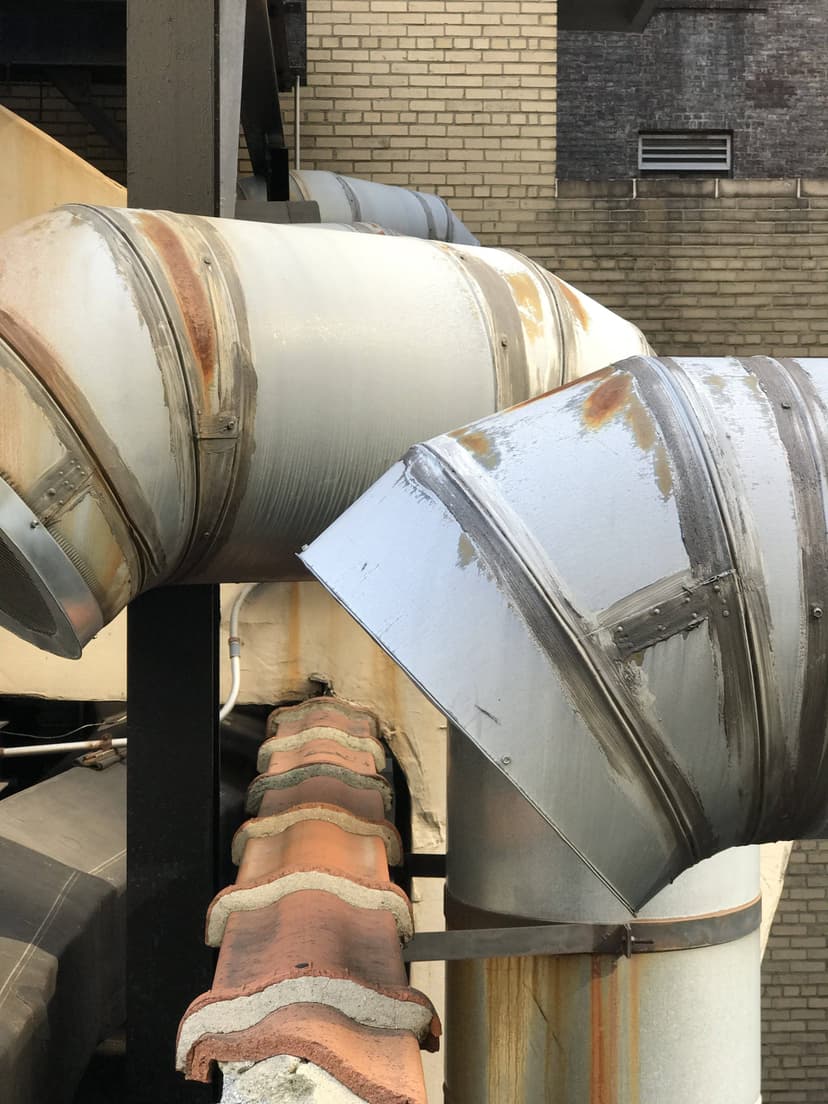 Rusty metal ducts bend and connect on a building exterior, with brick walls and ventilation in the background, showcasing a classic duct work example in HVAC systems.