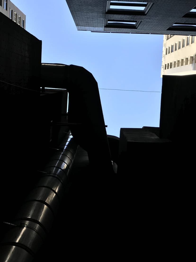 Looking up from the base of tall, dark buildings with gleaming HVAC duct work visible against the clear, blue sky between them.