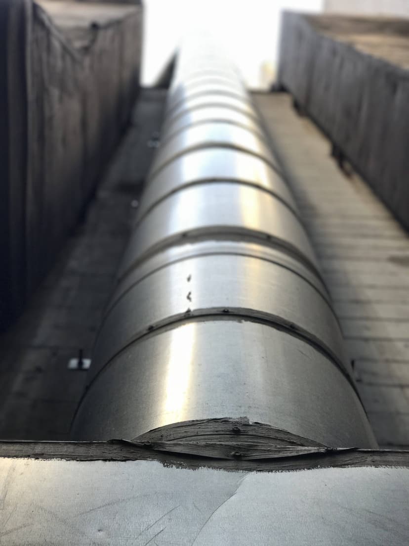 Looking up at a tall, silver metal pipe between two dark, narrow concrete walls, you see a prime example of intricate HVAC duct work.