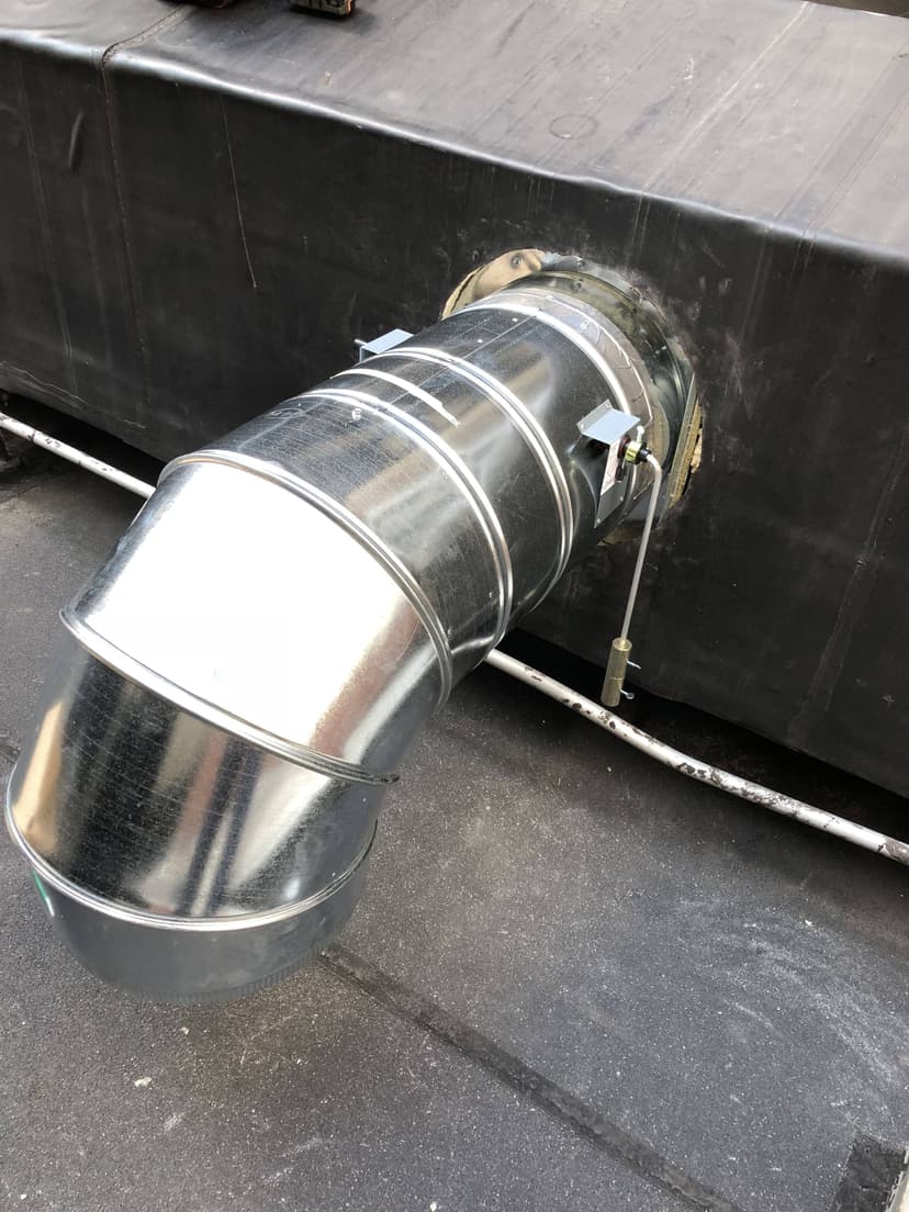 A large, metallic duct pipe serves as a prime HVAC example, attached at an angle to an exterior black wall on a rooftop.