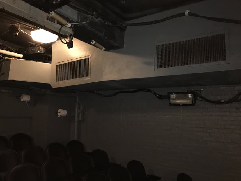 Dimly lit theater with black chairs, air vents, and exposed wiring on the dark gray walls, showcasing an example of intricate HVAC duct work.