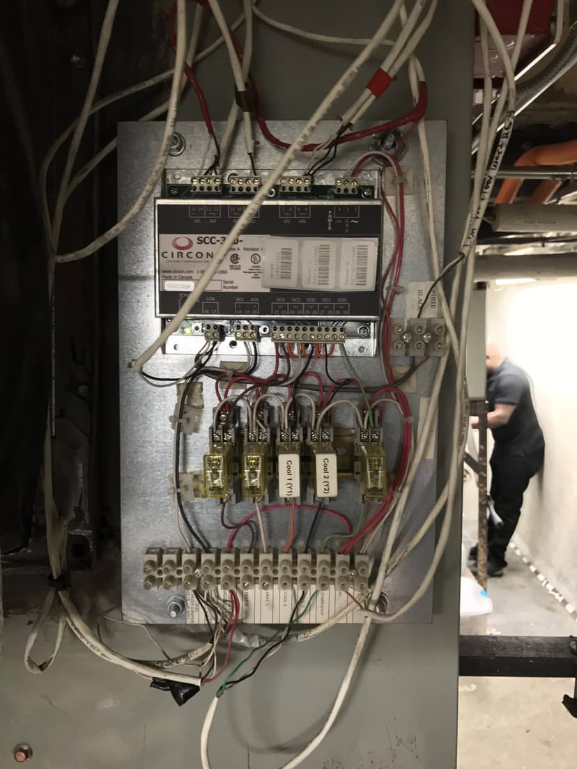 An open electrical control panel with various wires connected, likely part of an HVAC electric controls system; a person in the background.