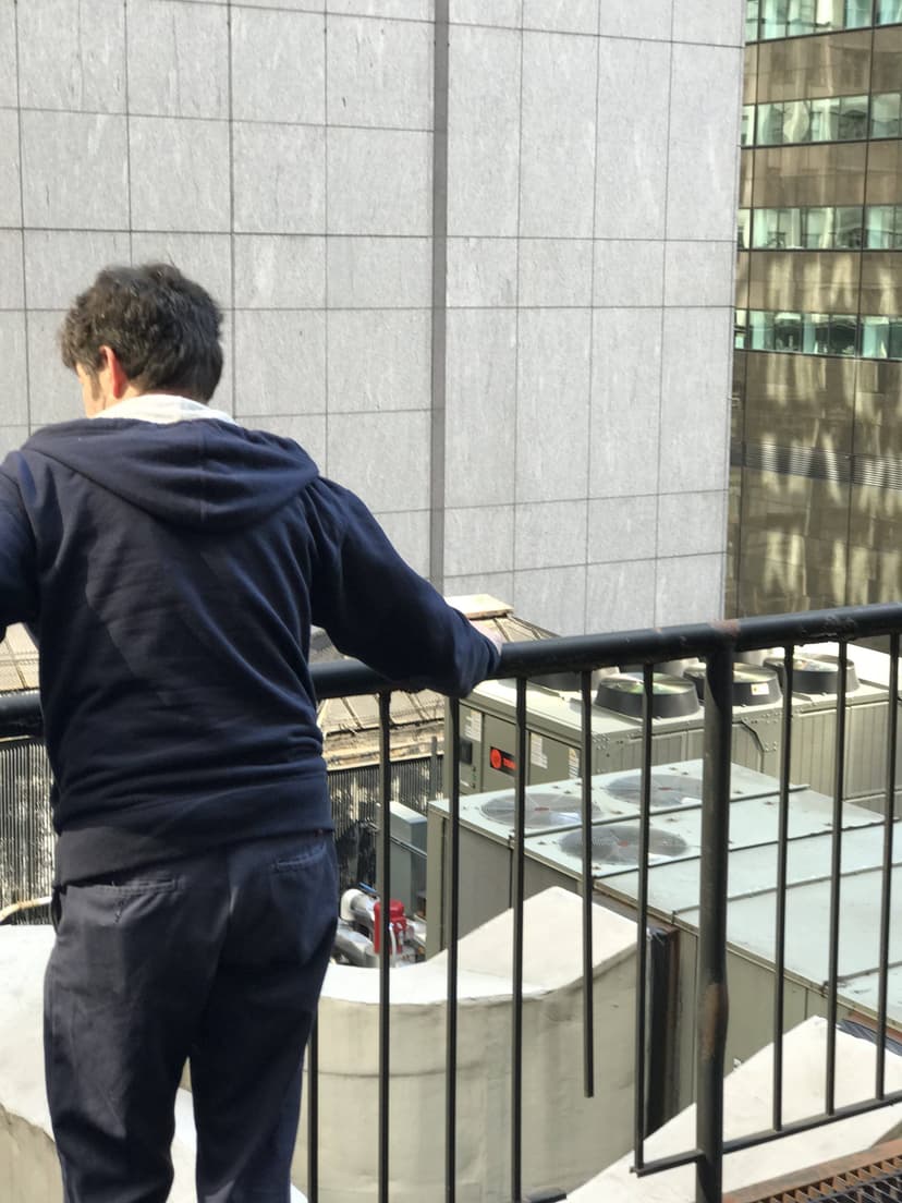 A person in a dark hoodie leans on a railing, looking out at industrial units and tall buildings in the cityscape, where HVAC mechanics keep NYC comfortable year-round.