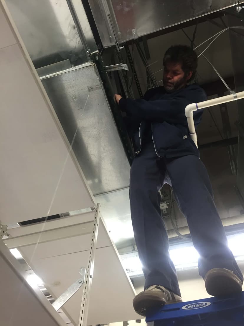 An HVAC mechanic in NYC is diligently working on the ceiling ventilation system, carefully balancing on a ladder in an industrial setting.