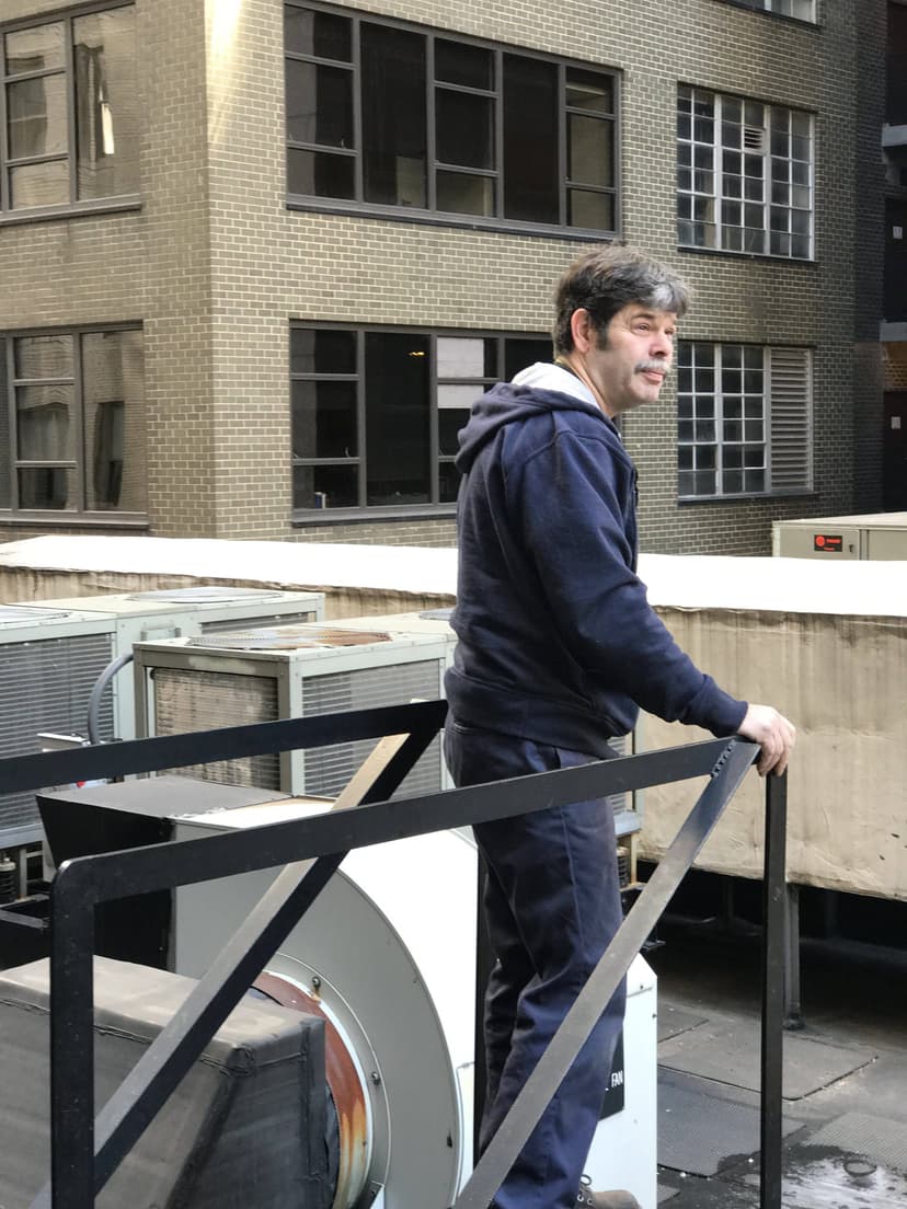 A man in a hoodie stands on a rooftop, holding a railing, with the sprawling NYC skyline and various HVAC units in the background.