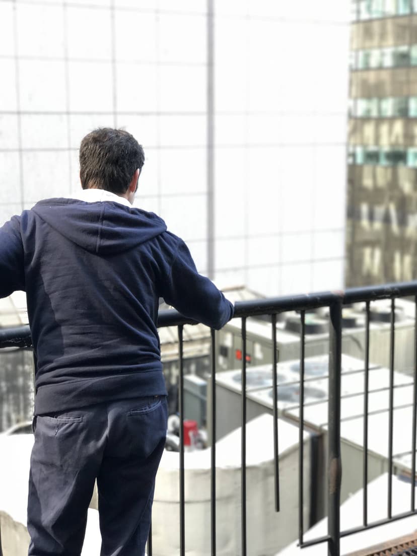 Person in a blue hoodie leaning on a railing, looking out over a cityscape with tall buildings and an overcast sky, where the intricate work of HVAC Mechanics NYC keeps every structure comfortable despite the weather.