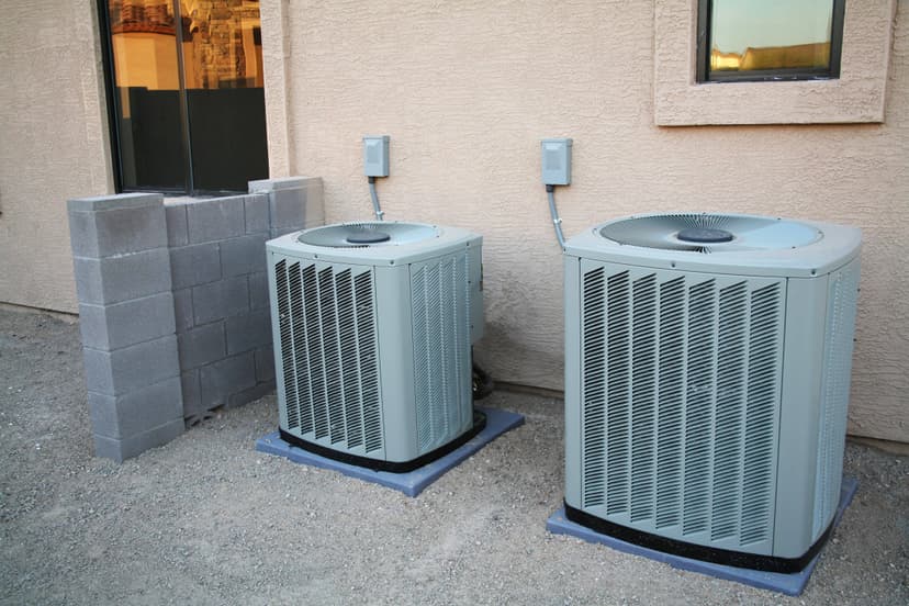 Two commercial HVAC systems are installed next to a beige stucco house, with a small cinder block wall nearby.