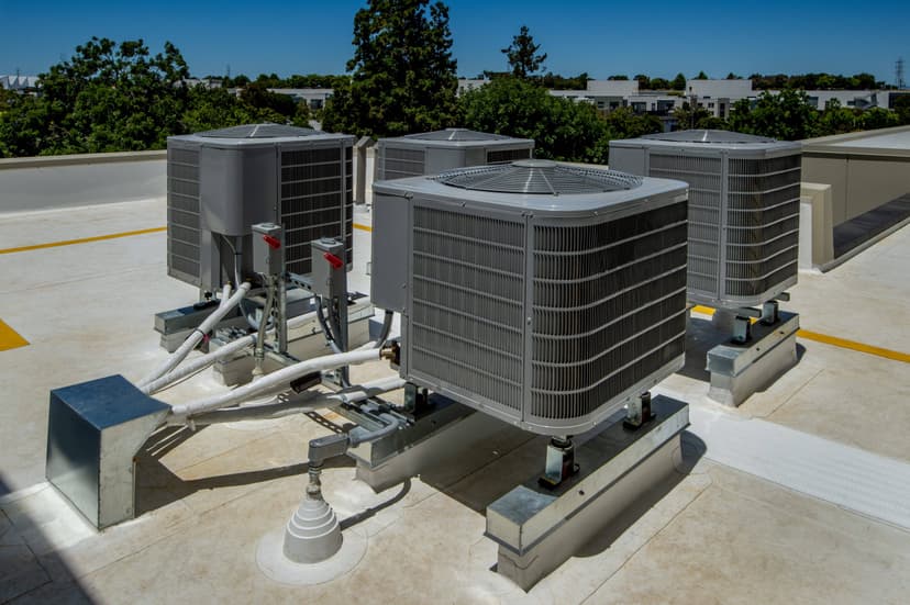 Four large commercial HVAC systems on a rooftop, surrounded by equipment and piping, gleam under the sunny sky.