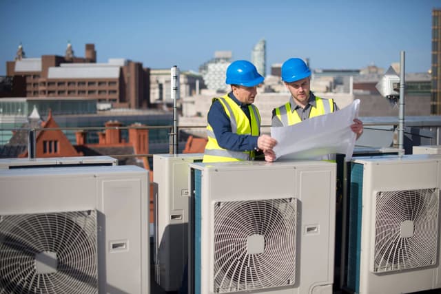 Two men standing on top of an air conditioning unit.