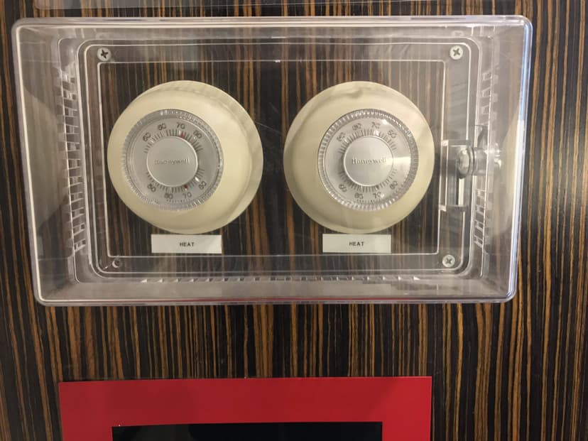 Two round Honeywell thermostats labeled Heat, part of the indoor HVAC system, are enclosed in a clear plastic cover, mounted on a wood-paneled wall. This arrangement exemplifies reliable commercial equipment.