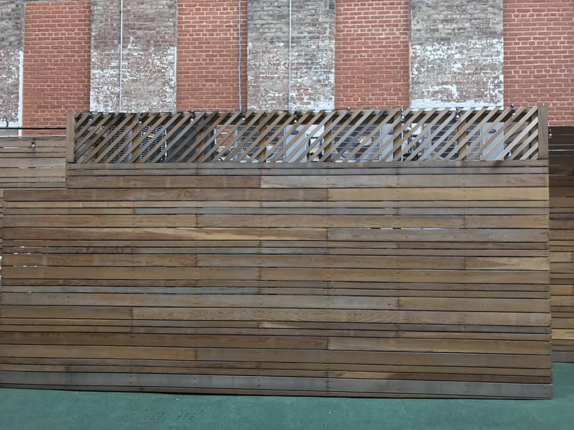 A wooden partition with a diagonal slat design at the top, set against a wall with red and white brickwork, discreetly conceals indoor commercial equipment.