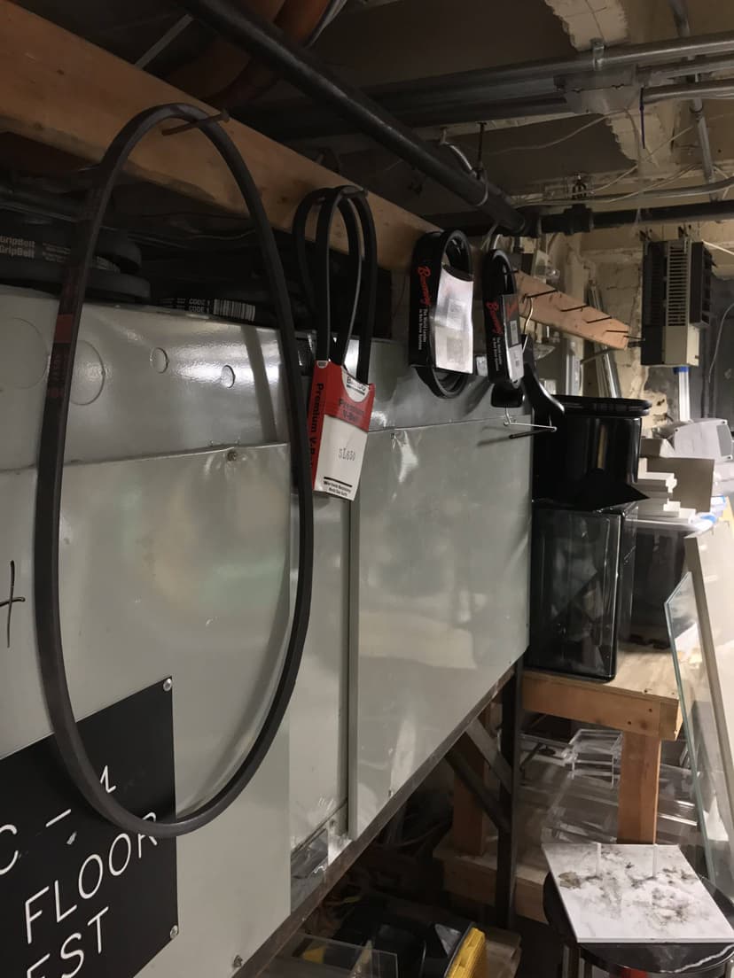 Industrial storage area with hanging belts, HVAC systems, electrical equipment, and various tools and supplies on shelves.