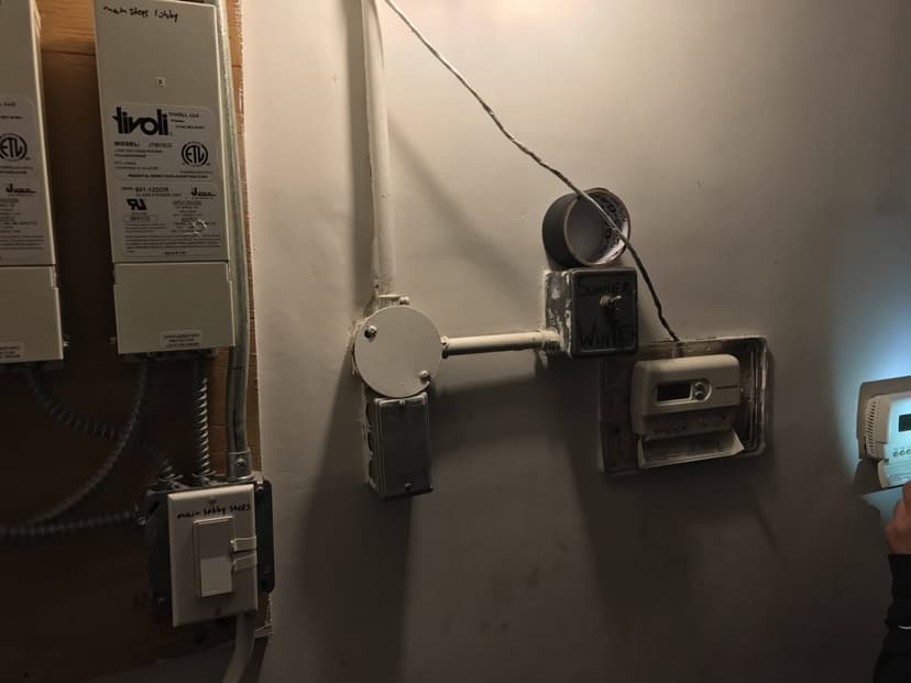 Electrical outlets, switches, and meters on a wall with wiring and duct tape, partially obscured person on the right, and indoor HVAC commercial equipment in the background.