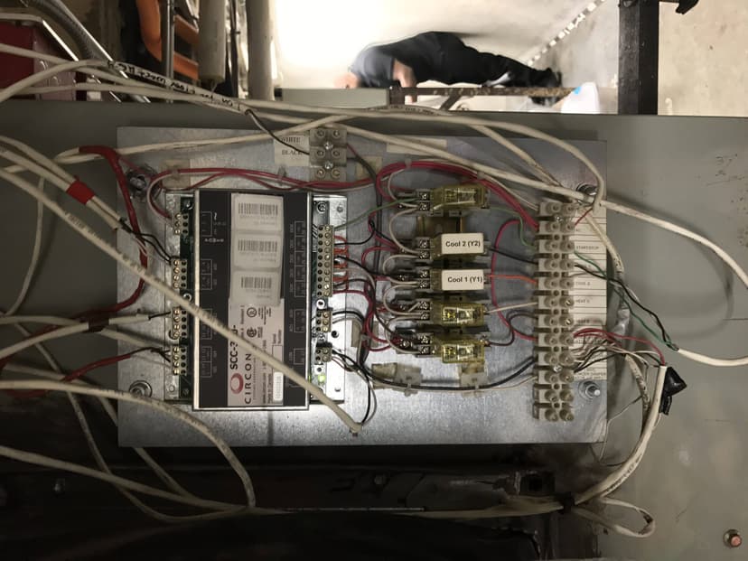 A control panel with wires and fuses is installed on a wall, part of the indoor HVAC commercial equipment setup, with blurred figures visible in the background.