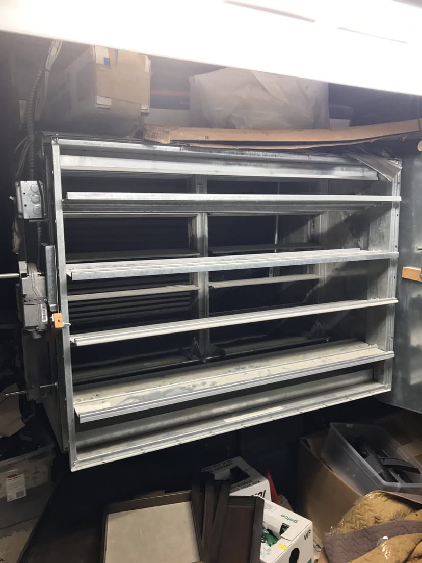 Large industrial vent with metal slats in a storage room, surrounded by boxes and miscellaneous commercial equipment.